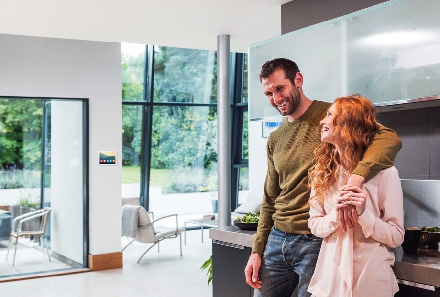 A happy couple standing in a modern, open-concept kitchen. The man is smiling and embracing the woman, who has long, curly red hair. They are surrounded by a bright and spacious room with large windows overlooking a lush, natural setting outside. 
