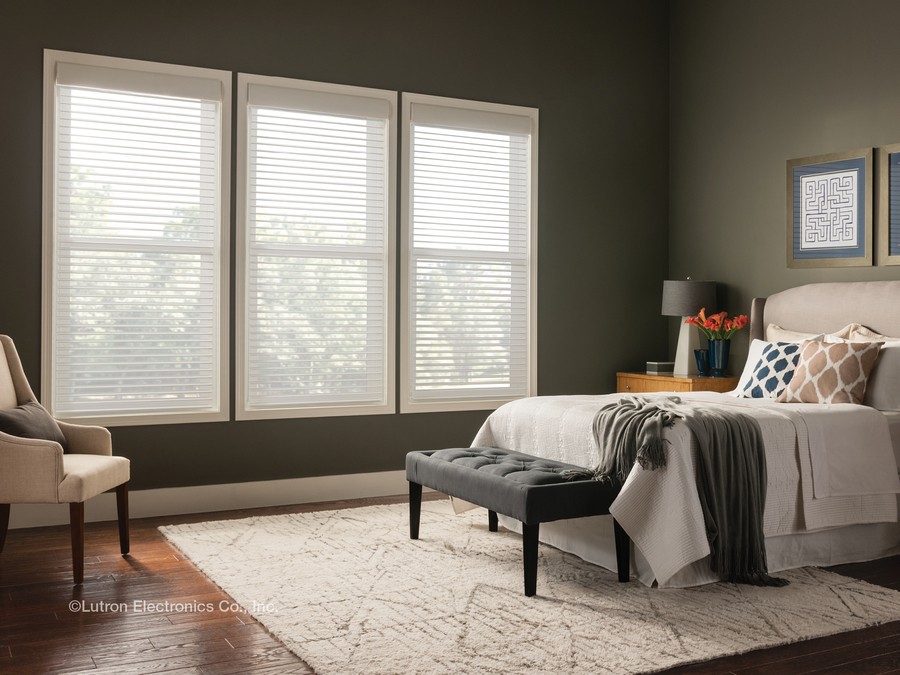 A bedroom with a bed with blankets draped over it and a chair facing it with light filtering in through sheer shades. 