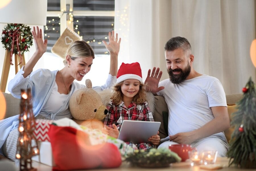 Two parents with their young daughter in between them on a couch with a window and shading behind them and holiday decor around them.